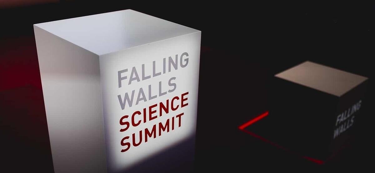 Photo of a white block on a dark stage with red and grey letters spelling out "Falling Walls Science Summit"