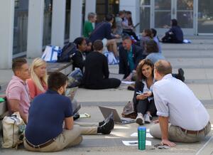 Groups of people sitting outside on the pavement, in discussion with each other, with papers strewn about. 